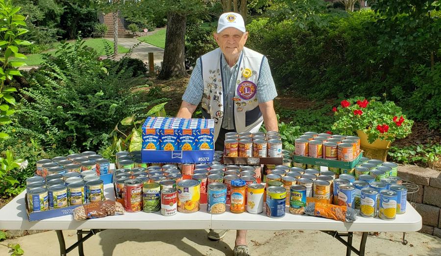 A Lexington Lions Club member in club cap and vest standing behind a table piled high with canned goods collected for a district food drive.