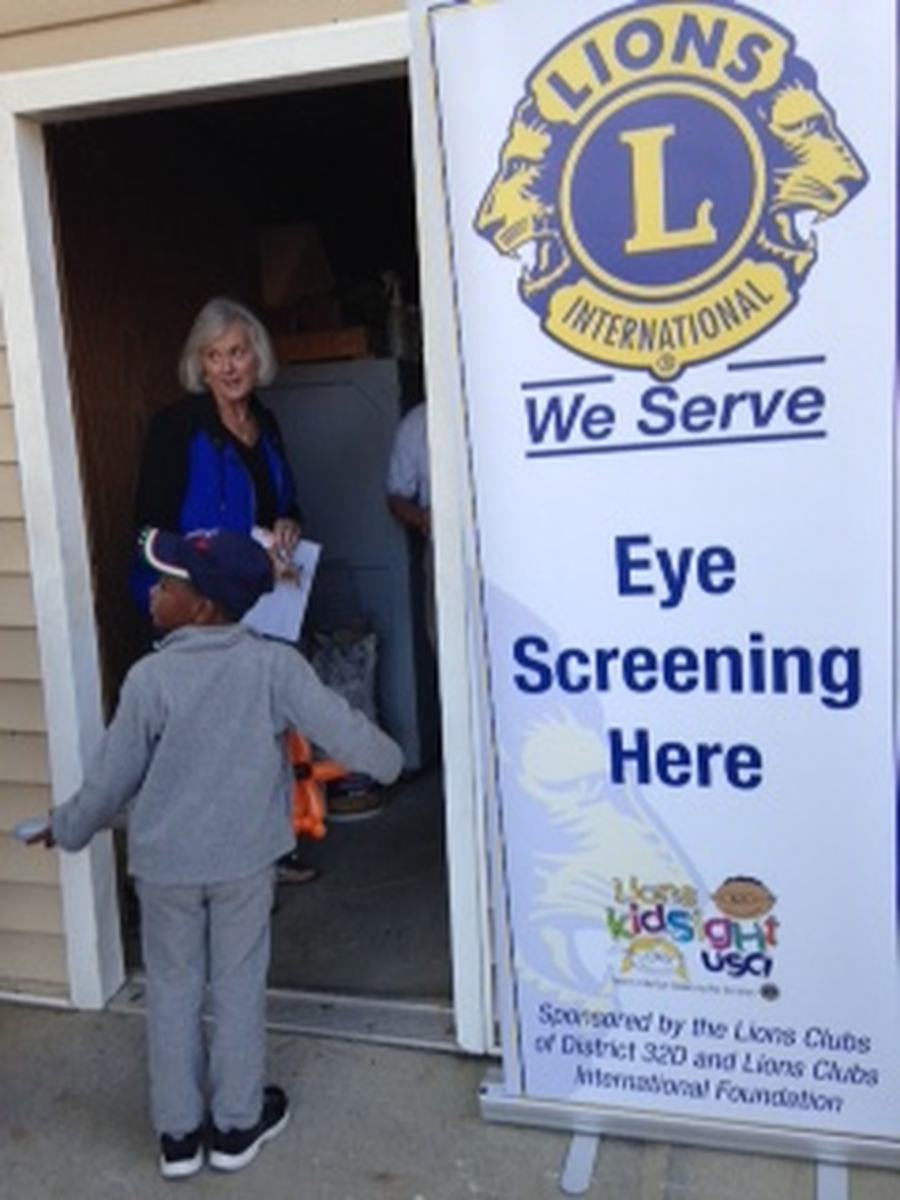 A Lexington Lions Club volunteer greets a child beneath a 'We Serve, Eye Screening Here' banner at a community vision-screening event.